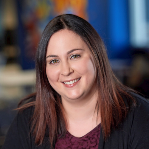 Smiling woman with straight, shoulder-length dark brown hair wearing a dark cardigan, photographed indoors with a blurred background.