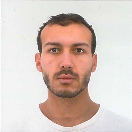 A man looking directly at the camera with a neutral expression. He has short dark hair, light stubble, and is wearing a plain white crew-neck shirt against a solid white backdrop.