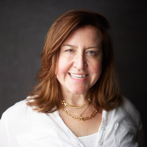 Smiling woman with shoulder-length auburn hair wearing a white blouse and layered gold necklaces against a dark background.