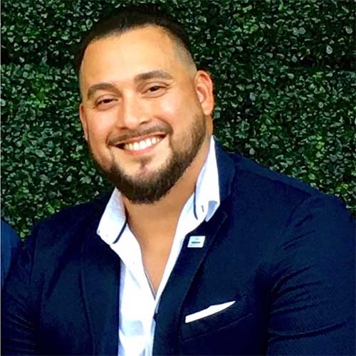 Smiling man with short dark hair and a trimmed beard wearing a navy blazer and open-collar white shirt in front of a green leafy backdrop.