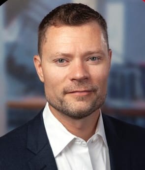 Man with short brown hair and a trimmed beard, wearing a navy blazer and white button-down shirt, facing the camera against a softly blurred office background.
