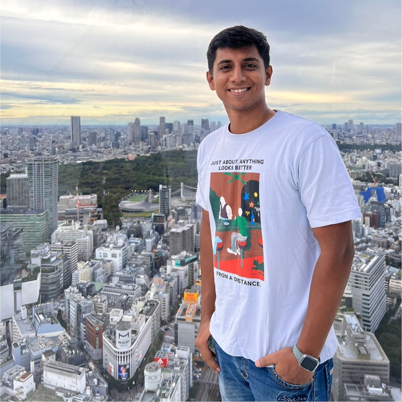 Smiling man in a white graphic T-shirt and jeans standing on front of a panoramic city skyline from a high-rise viewpoint.
