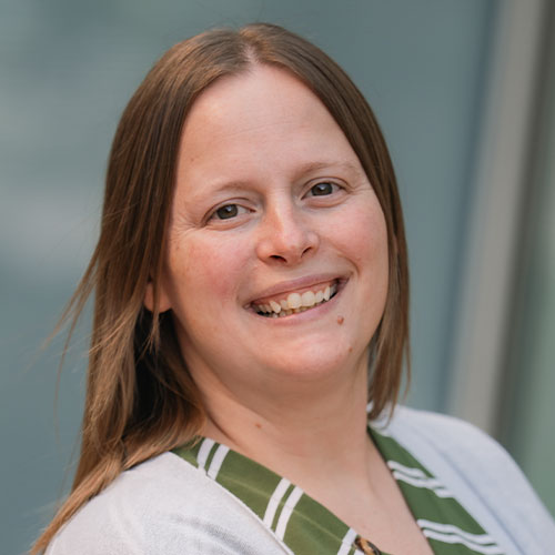 Smiling woman with long light brown hair, wearing a green striped blouse and light cardigan, facing the camera against a blurred blue-gray background.