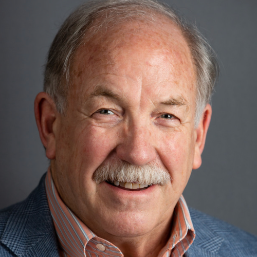 Smiling man with gray hair and a gray mustache in a close-up headshot against a gray background.