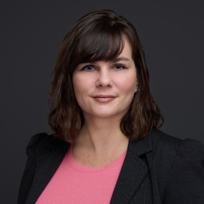 Professional headshot of a woman with shoulder-length dark brown hair wearing a black blazer and pink top against a dark gray background.