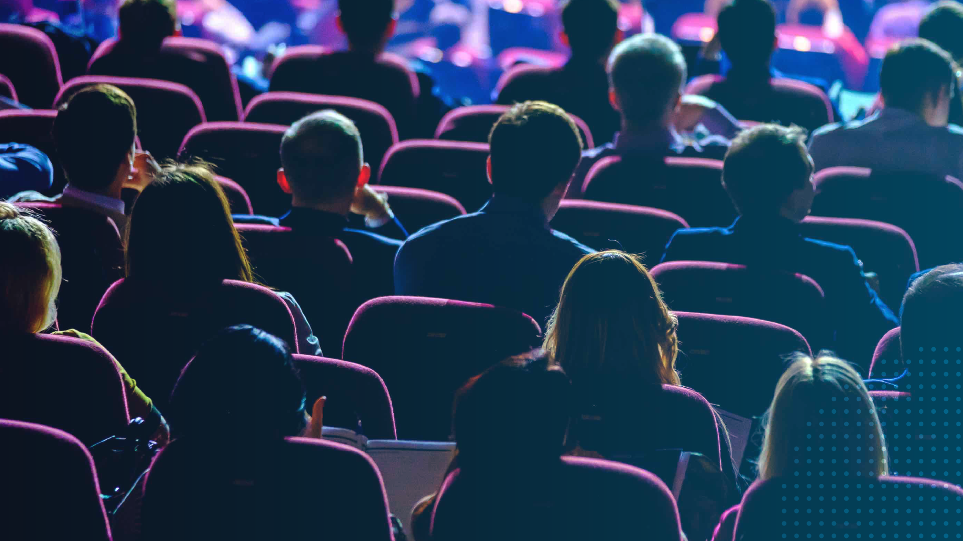 Audience members seated in rows at a large conference or keynote session, viewed from behind, with attendees listening attentively in a dimly lit auditorium.