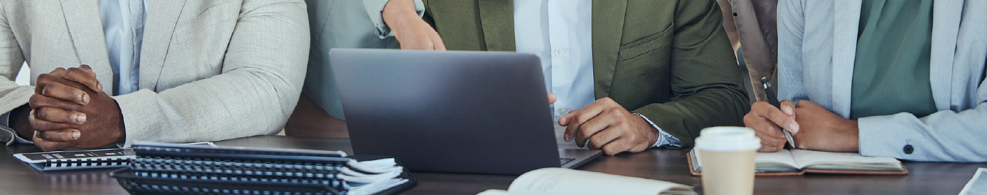 Three professionals collaborating around a laptop at a table with notebooks and coffee.