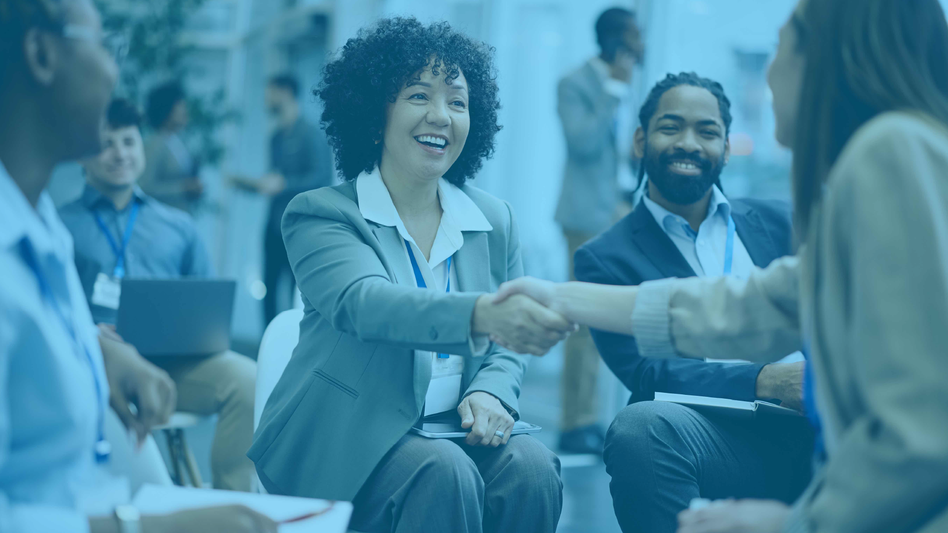 Attendees shaking hands and smiling during a small-group networking discussion at an MRC Connects regional event, with other professionals conversing in the background.