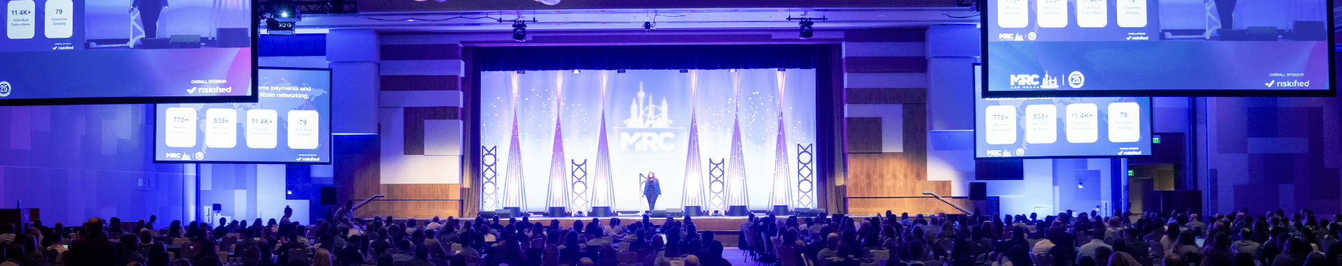 Wide-angle view of a packed conference hall during an MRC Vegas keynote presentation, with a speaker on stage under bright lights and large screens displaying event statistics and the Riskified sponsor logo.