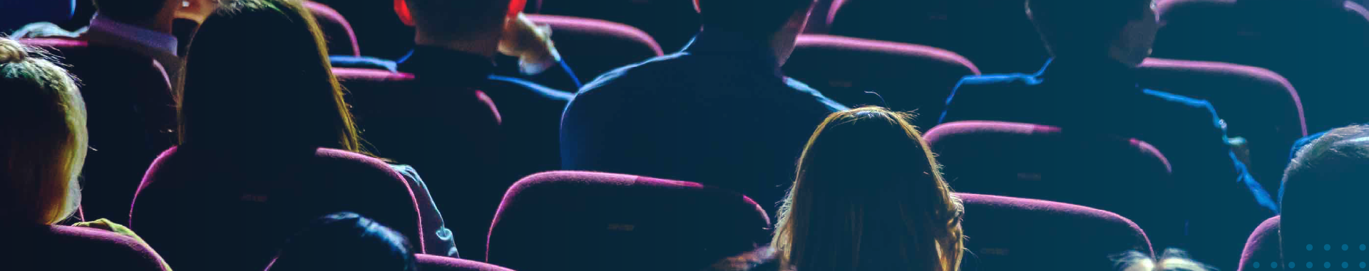Conference crowd seen from behind in purple auditorium seats, focused on a brightly lit stage with blue-purple lighting.
