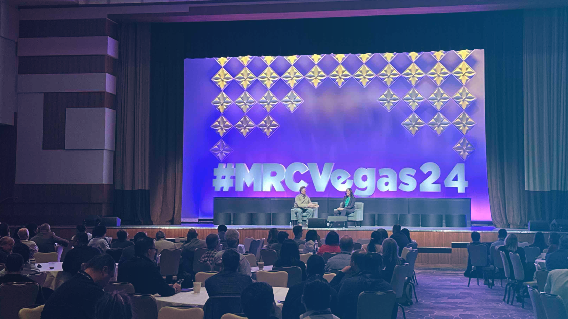 Conference audience seated at round tables watching two speakers in armchairs on stage at MRC Vegas 2024. The stage backdrop features large illuminated text reading ‘#MRCVegas24’ with a geometric diamond pattern above.