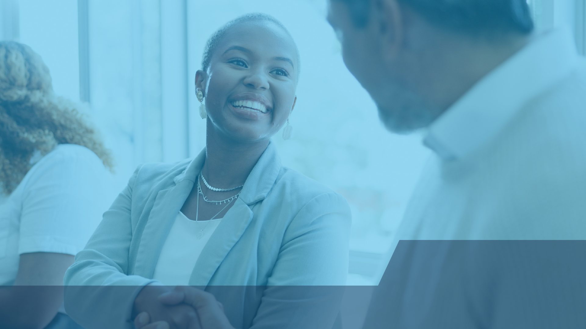 Two professionals smiling and shaking hands during a one-on-one conversation at an MRC Connects networking event, with other attendees engaged in discussion nearby.