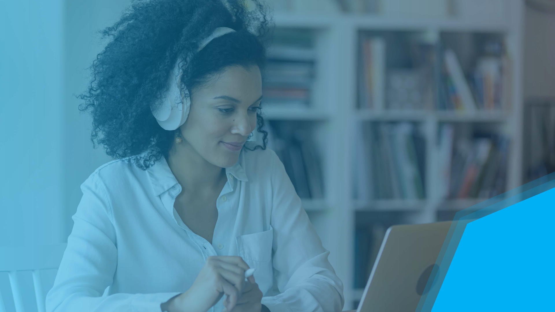 Person wearing headphones smiles while working on a laptop at a desk, with bookshelves in the background.