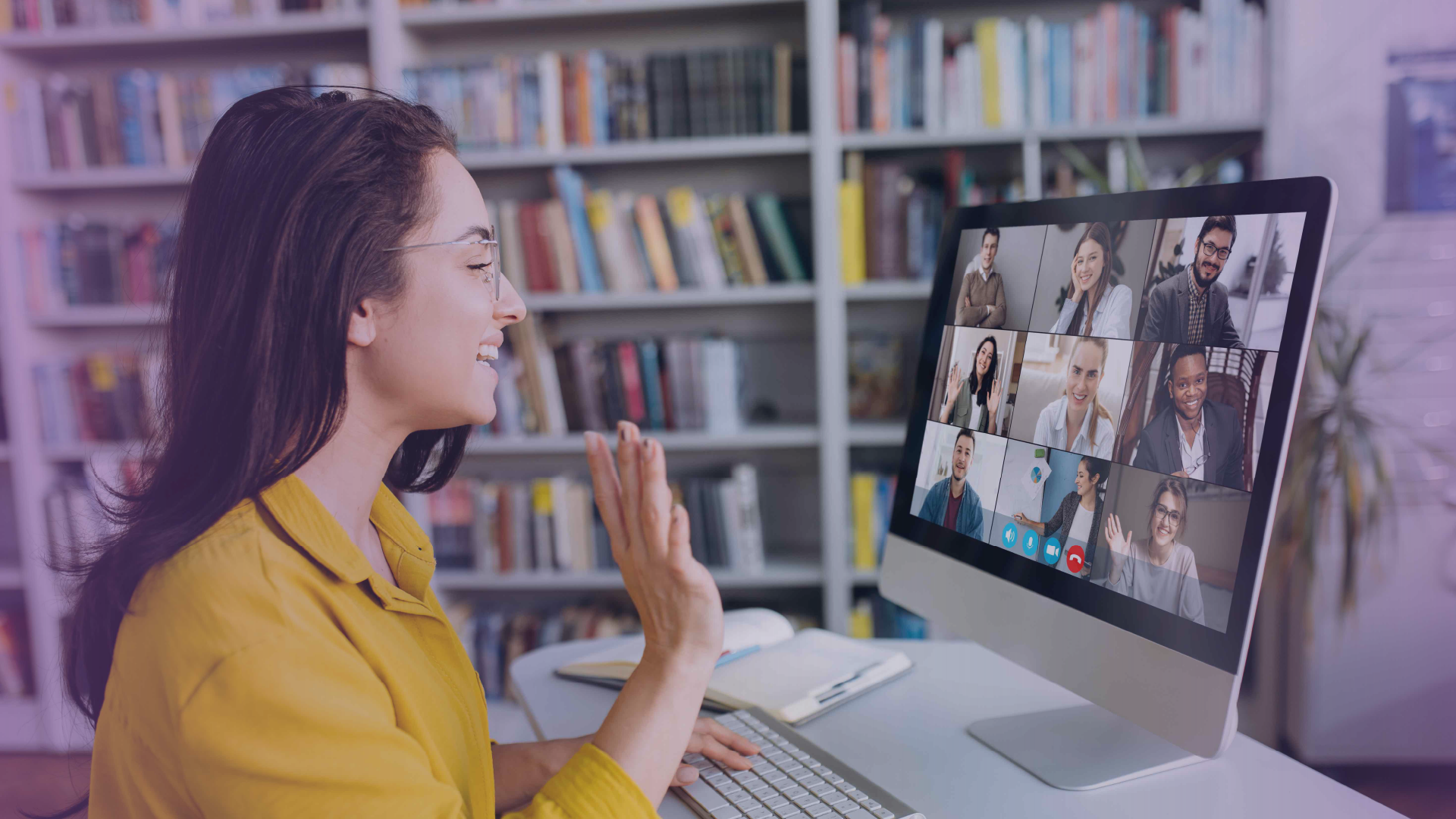 Professional waving and engaging in a virtual summit from a desk, with multiple attendees visible on a video call screen in a home office setting.