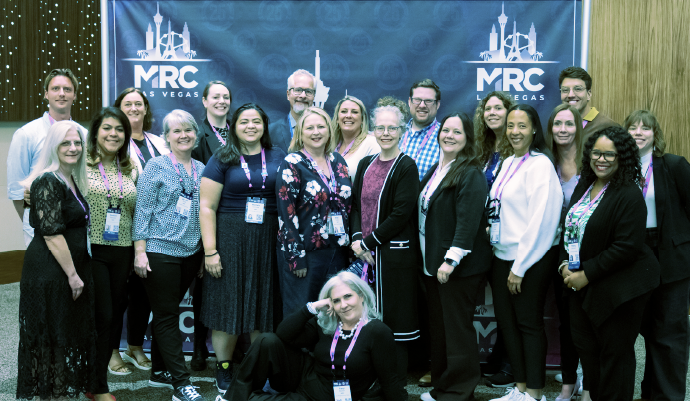 Group photo of MRC staff and attendees standing and smiling in front of a branded MRC Vegas backdrop at a conference event, wearing name badges and business casual attire.